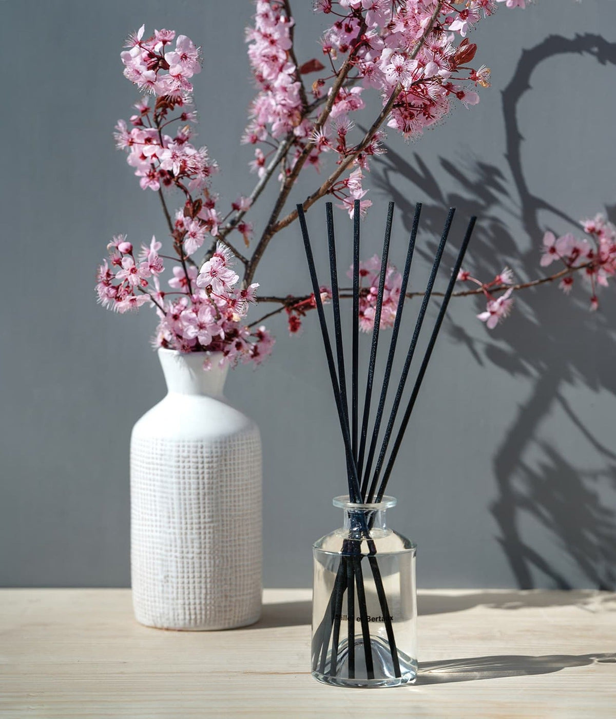 Miller et Bertaux Diffusor In The Garden Refill neben einer weißen Vase mit blühenden Kirschzweigen