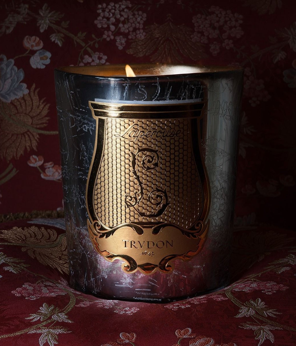 Trudon Duftkerze Maison de Plaisirs brennend auf rotem Damast mit Blumenmuster